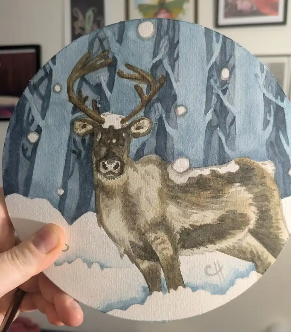 A hand holds up a circular watercolor painting of a reindeer standing in a snowy forest with bare trees and falling…
