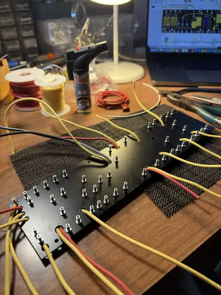 A cluttered electronics workbench features a black circuit board with numerous metal pins and colorful wires (yellow, red…