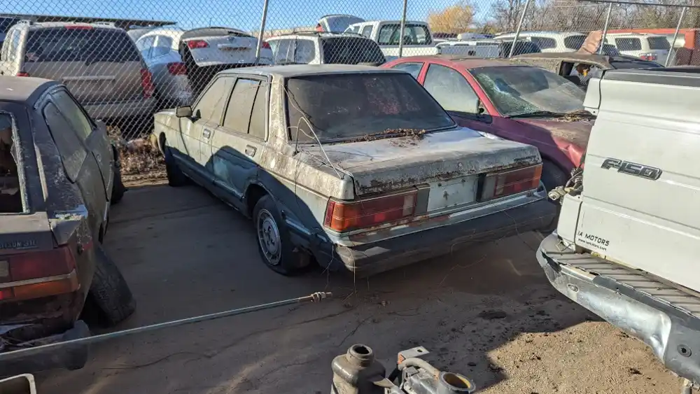 A heavily rusted, dirty, and abandoned sedan sits in a junkyard surrounded by other derelict vehicles. The car's rear is…