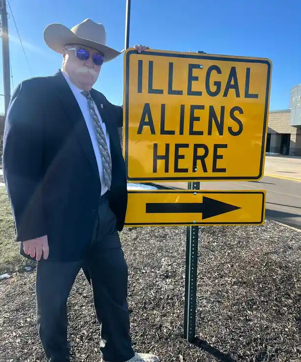A man in a cowboy hat, sunglasses, and suit stands next to a yellow road sign that reads 'ILLEGAL ALIENS HERE' with an arrow…
