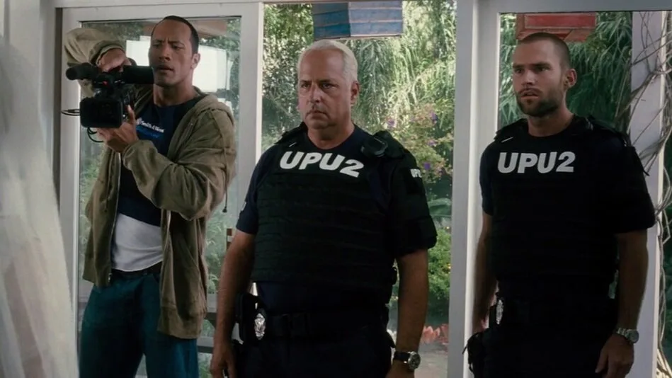 Three men stand in a doorway; one holds a camera, two wear black tactical vests with 'UPU2' printed in white. The scene is…
