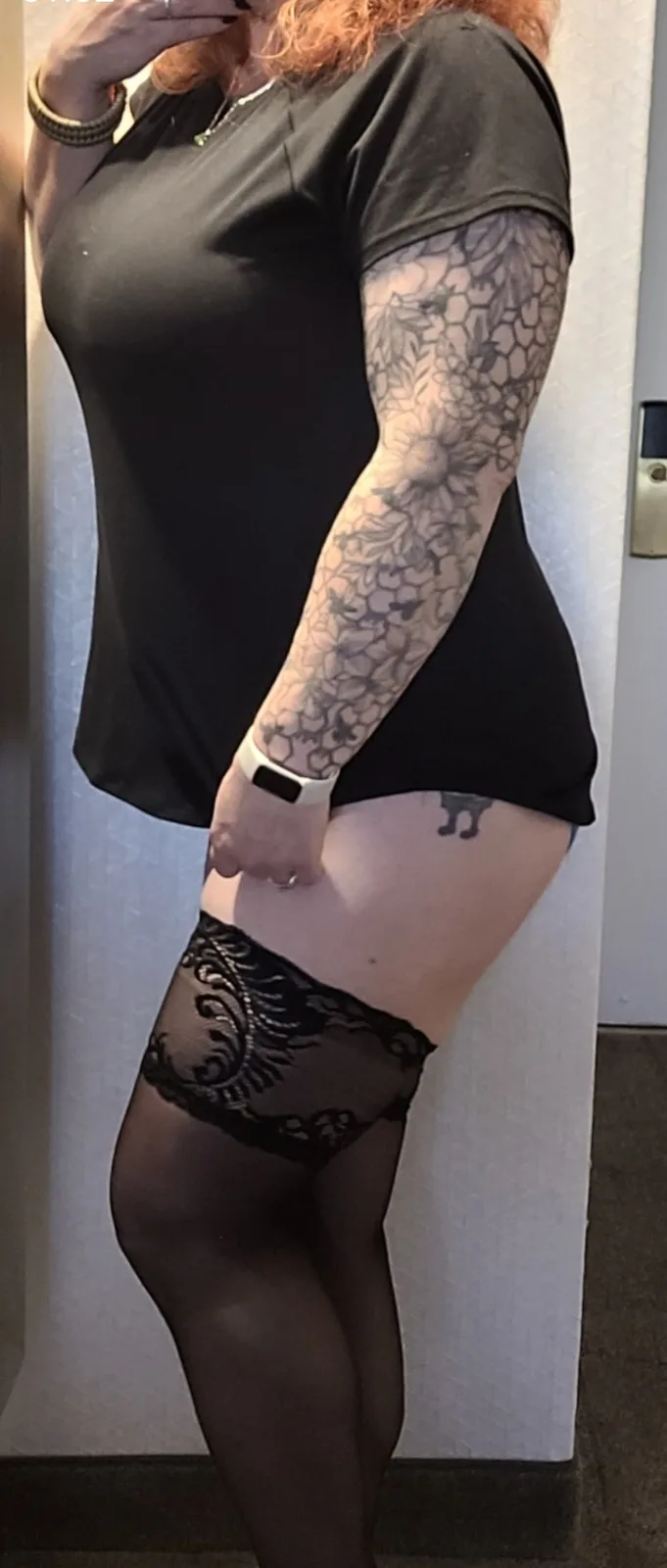 A person with red hair and a black t-shirt stands in front of a mirror, showing off a large floral tattoo on their left arm…