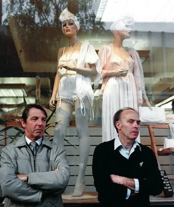 Two men stand in front of a store window with two female mannequins dressed in white lingerie and head coverings. Both men…