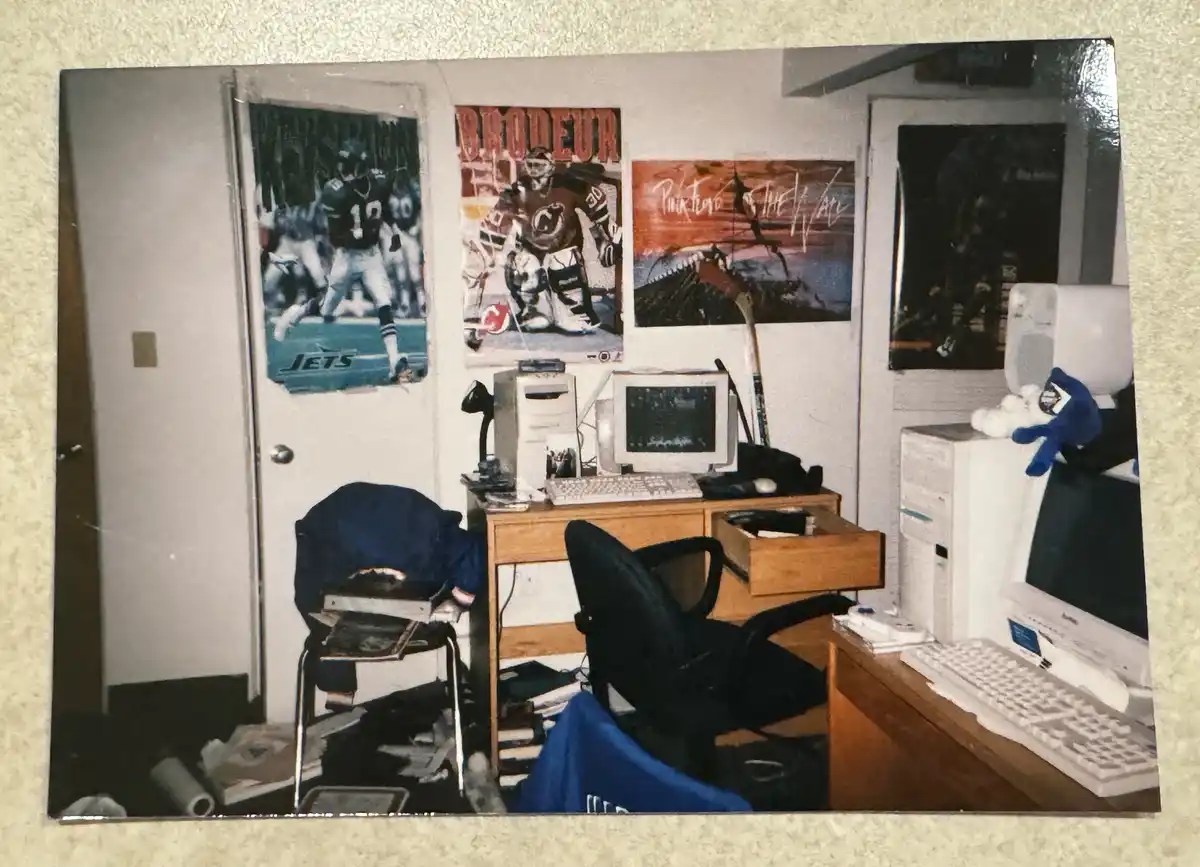 A cluttered, nostalgic bedroom photo shows a wooden desk with two old white computer setups, a black office chair, and…