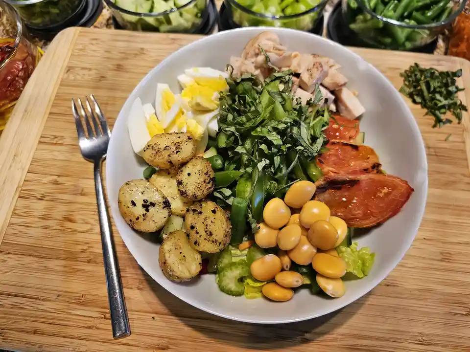 A white bowl filled with a colorful, neatly arranged salad sits on a wooden cutting board, containing roasted potatoes…
