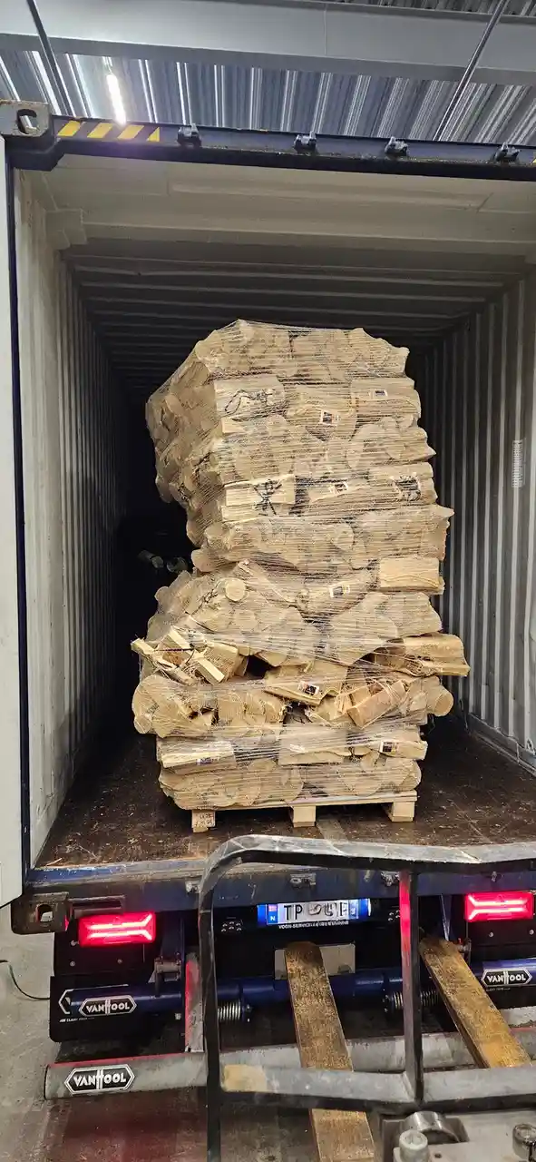 A large, tightly packed stack of firewood wrapped in netting sits inside an open shipping container, viewed from the back of…