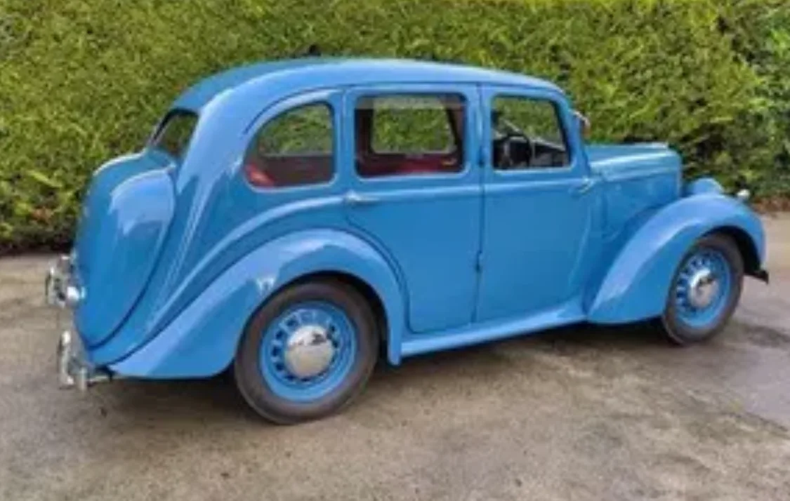 A vintage blue sedan with rounded fenders and chrome hubcaps is parked on a paved surface in front of a green hedge. The…