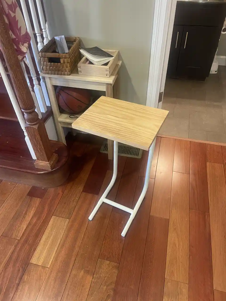 A simple, homemade wooden table with white metal legs sits on hardwood flooring near a staircase. In the background, a small…