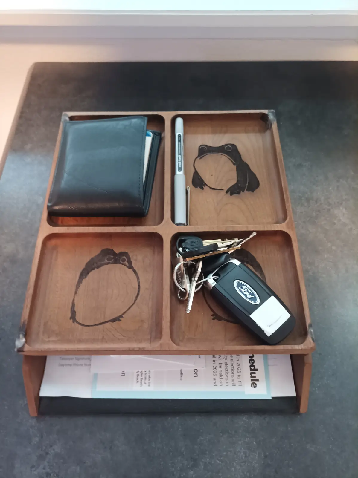 A wooden four-compartment tray holds a black wallet, a gray marker, a Ford car key fob with keys, and papers underneath…