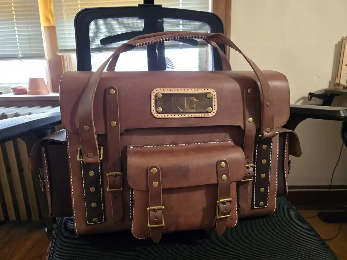 A brown leather satchel with brass hardware and white stitching sits on a black office chair, featuring a metal plaque with…