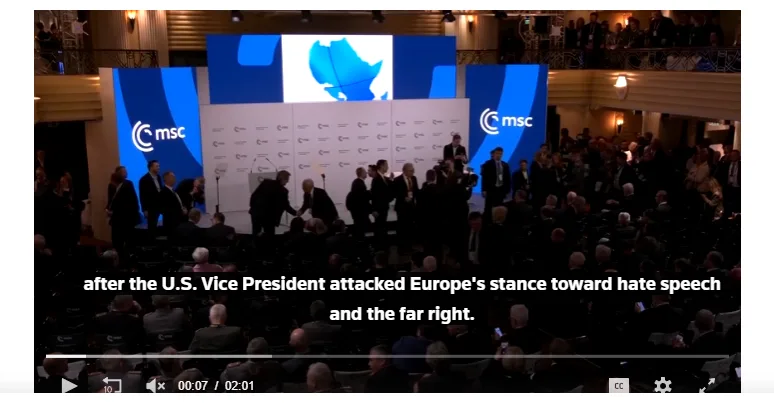 A screenshot from a video shows a formal conference with many people in suits on a stage in front of a large screen…