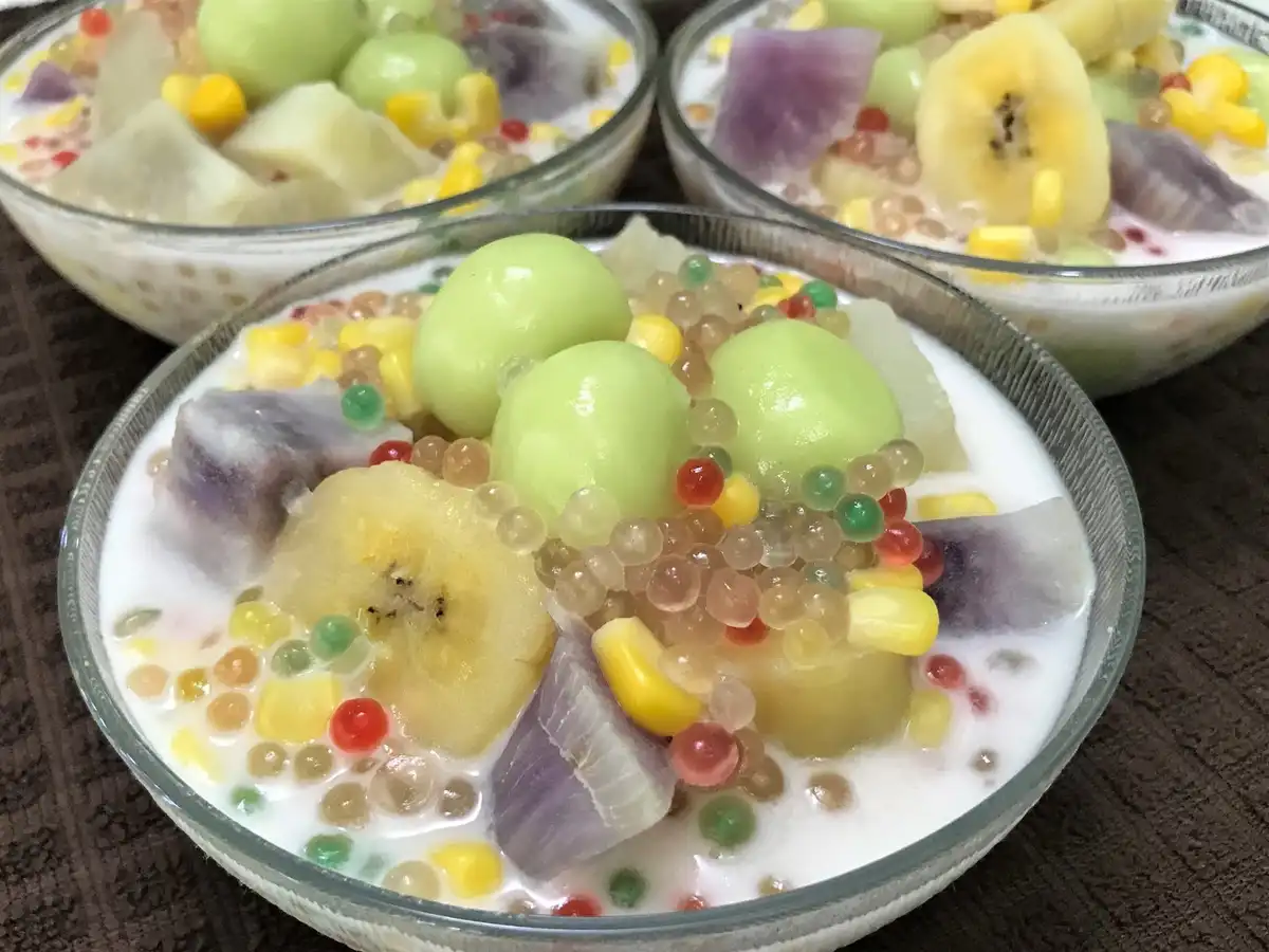 Three bowls of colorful Thai dessert, featuring sweet sticky rice balls, sliced bananas, purple yam, corn kernels, and…
