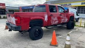 A red pickup truck is parked in a lot with visible damage and graffiti on its rear and side. The truck has large black tires…