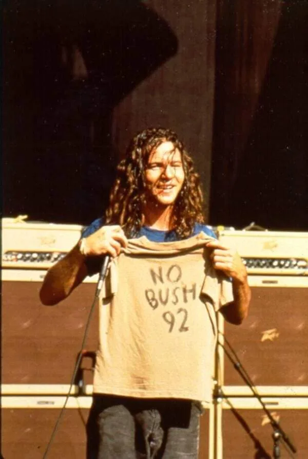 A man with long curly hair stands on a stage in front of amplifiers, holding up a beige t-shirt with the handwritten text…