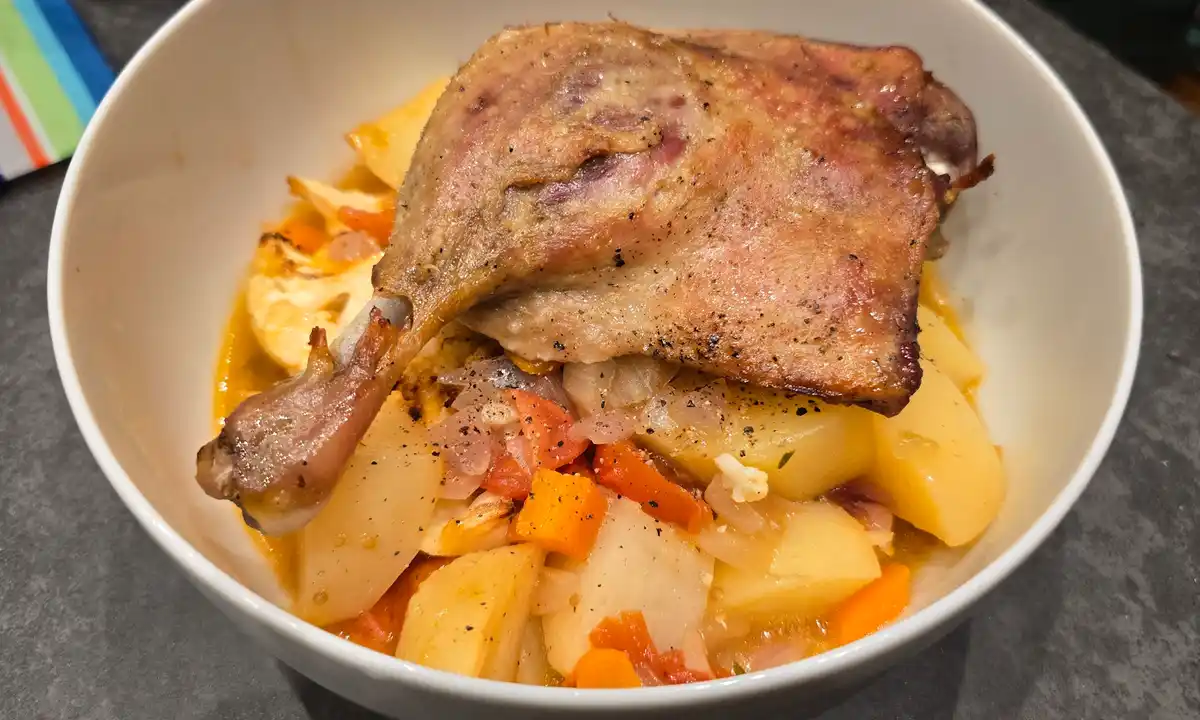A close-up photo shows a white bowl filled with a stew containing a large roasted duck leg, chunks of potato, carrots…