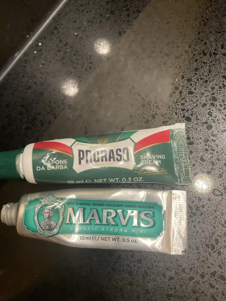 Two small tubes lie on a speckled countertop: a green Proraso shaving cream tube and a silver Marvis toothpaste tube, both…