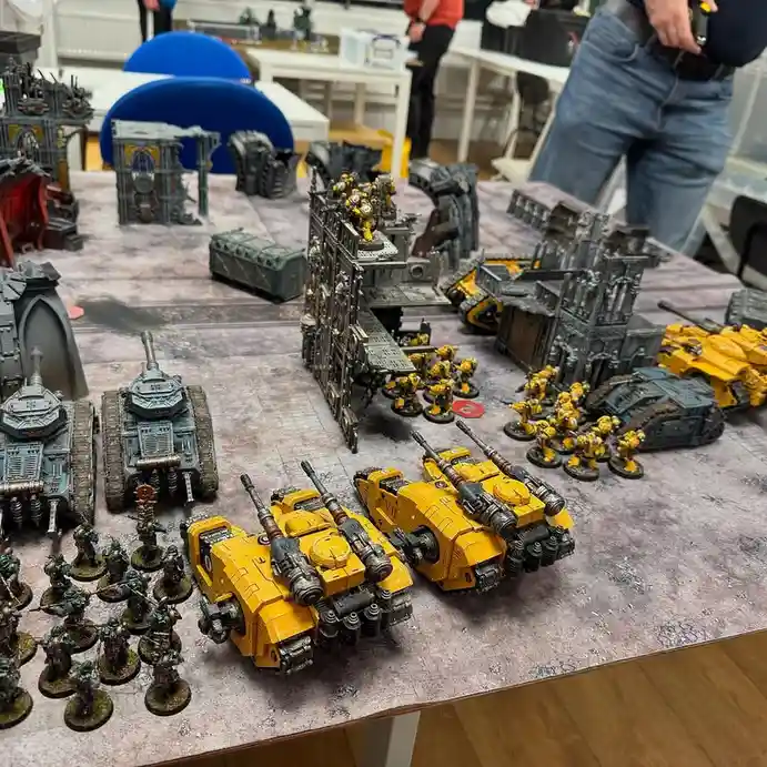 A tabletop wargaming scene shows two armies facing off: one side has numerous yellow-painted tanks and infantry, while the…