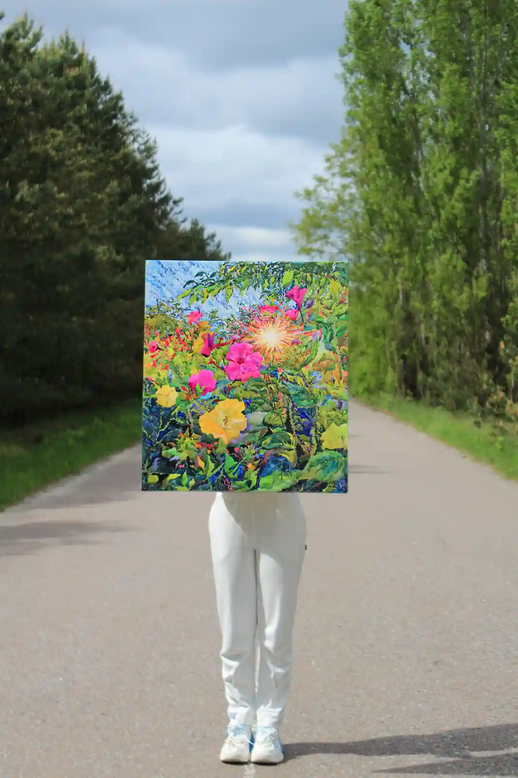 A person standing on a paved path between green trees holds a large, colorful painting of flowers and foliage in front of…