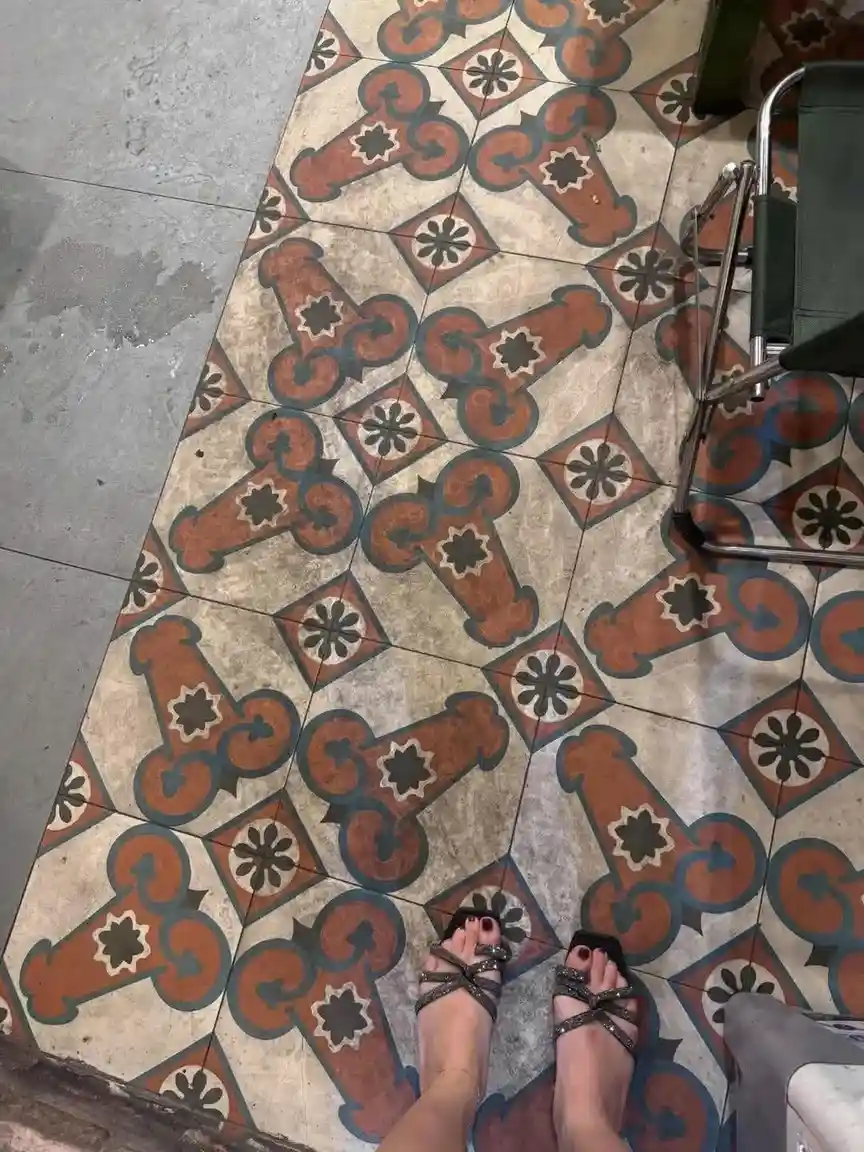 A first-person view shows a person's feet in sparkly black sandals standing on a patterned tile floor with orange, blue, and…