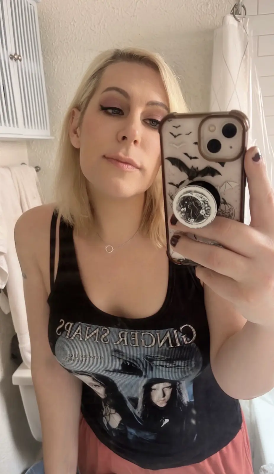 A woman with blonde hair takes a mirror selfie in a bathroom, wearing a black tank top featuring the movie 'Ginger Snaps'…