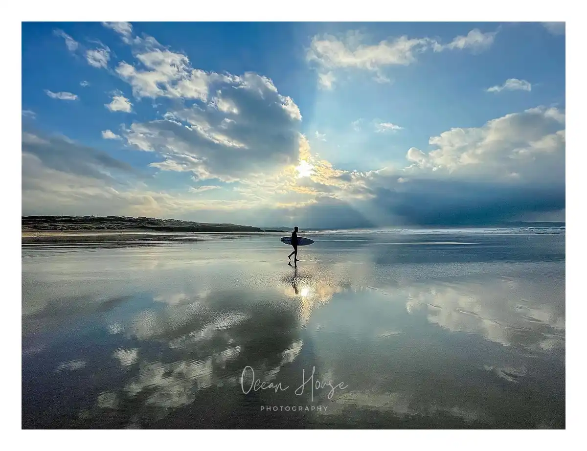A silhouetted surfer walks across a wet, reflective beach toward the ocean under a dramatic sky with sunbeams piercing…
