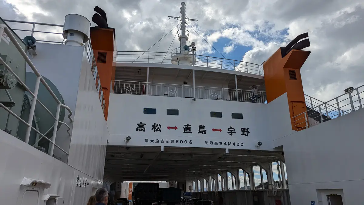 A low-angle photo of a large white ferry with orange funnels, showing its entrance and signage in Japanese. The text reads…