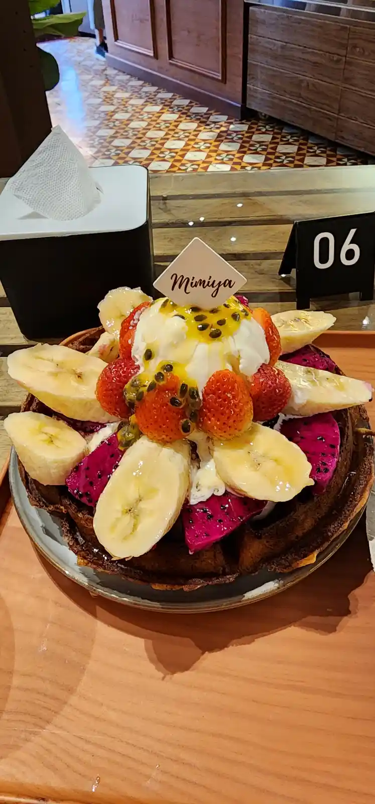 A close-up photo of a waffle topped with sliced bananas, strawberries, dragon fruit, whipped cream, and passion fruit seeds…