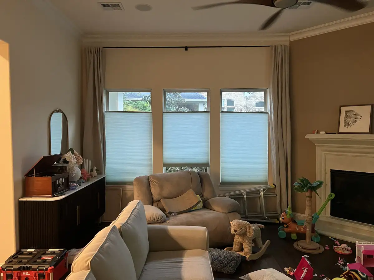 A cozy living room with beige sofas, a fireplace, and three windows with blinds, cluttered with children's toys like a…
