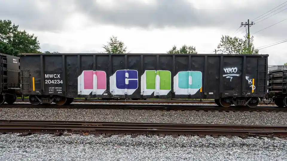 A black freight train car sits on tracks under a cloudy sky, featuring large, colorful graffiti that spells out 'DGO' in…
