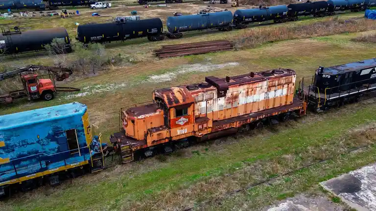 An aerial photo shows several rusty and abandoned railroad locomotives and tanker cars sitting idle on tracks in a grassy…