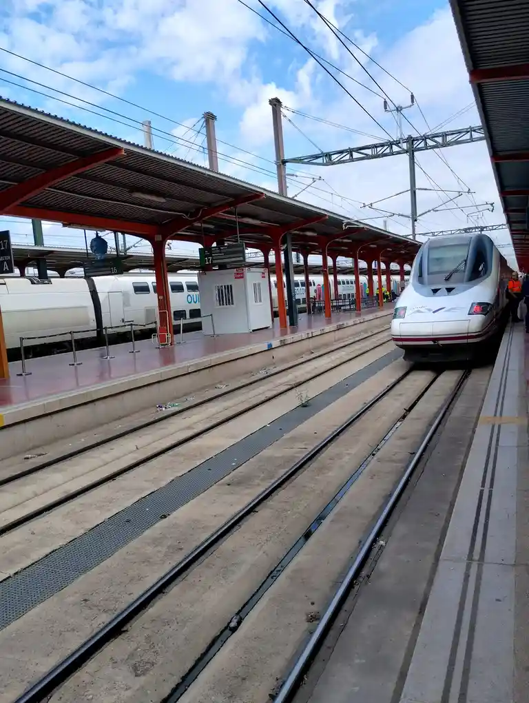 A high-speed train is stopped at a station platform, with another train visible on an adjacent track under a red-roofed…