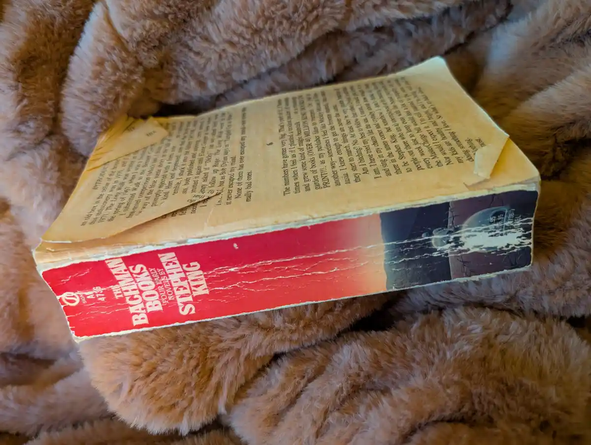 A worn, vintage paperback book titled 'The Bachman Books' by Stephen King rests on a fluffy brown blanket. The back cover…