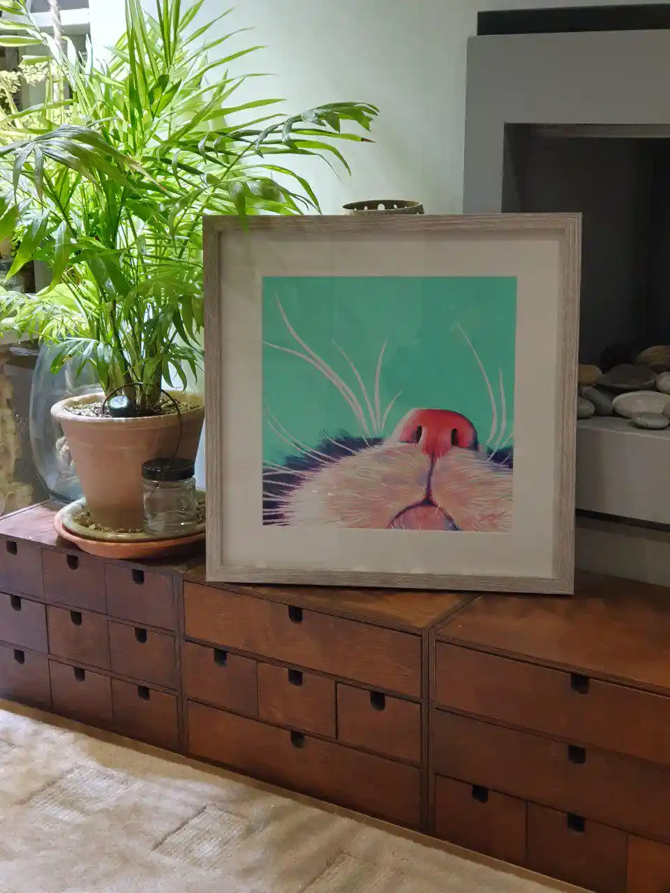 A framed painting of a cat's nose and whiskers against a teal background sits on a wooden dresser next to a potted plant…