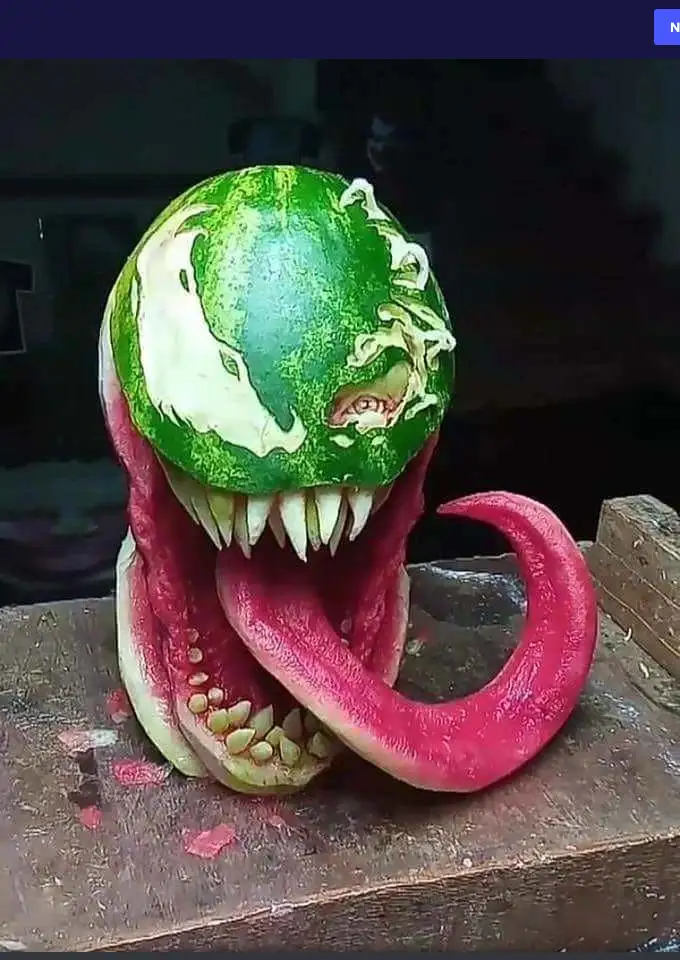 A watermelon has been intricately carved to resemble the comic book character Venom, featuring a green, textured head with…