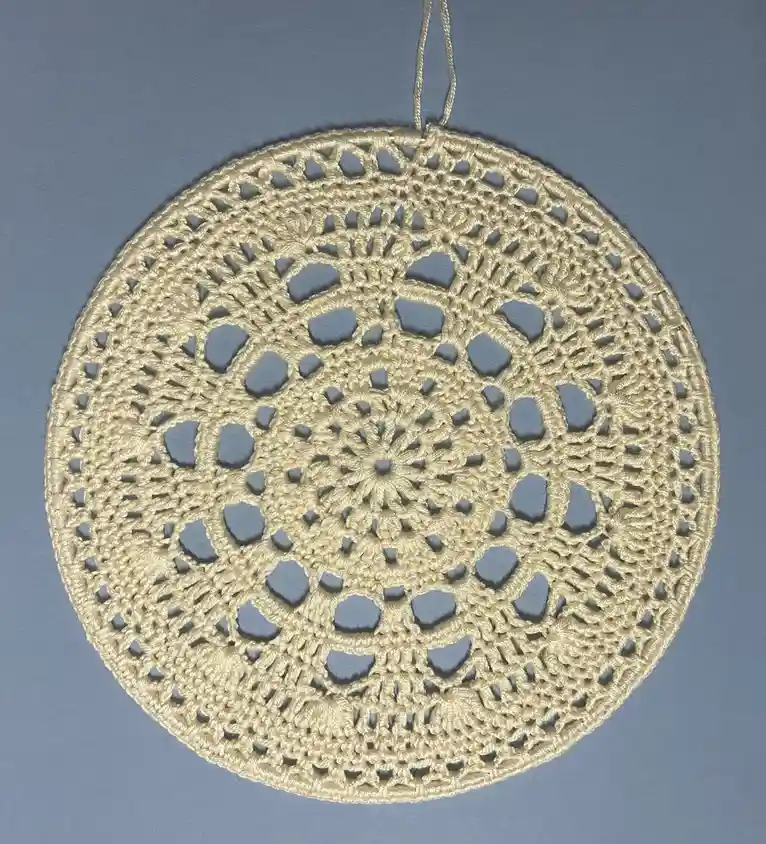 A circular, beige crochet doily with intricate lace patterns and a hanging loop is photographed against a solid blue-gray…