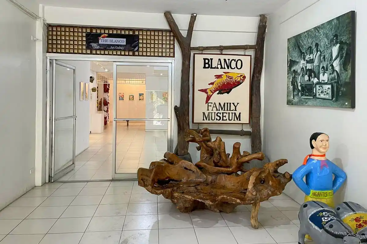 An indoor museum entrance with a large wooden root sculpture, a painted sign reading 'BLANCO FAMILY MUSEUM' with a fish…