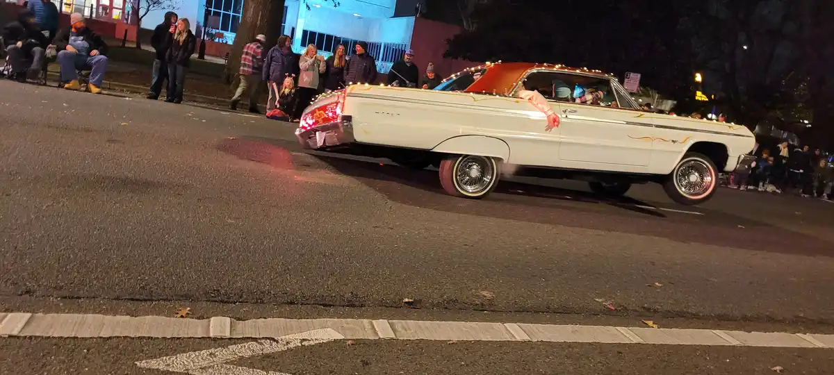 A white classic car with a red roof and decorative lights is shown at night, lifted on its front wheels as if performing a…