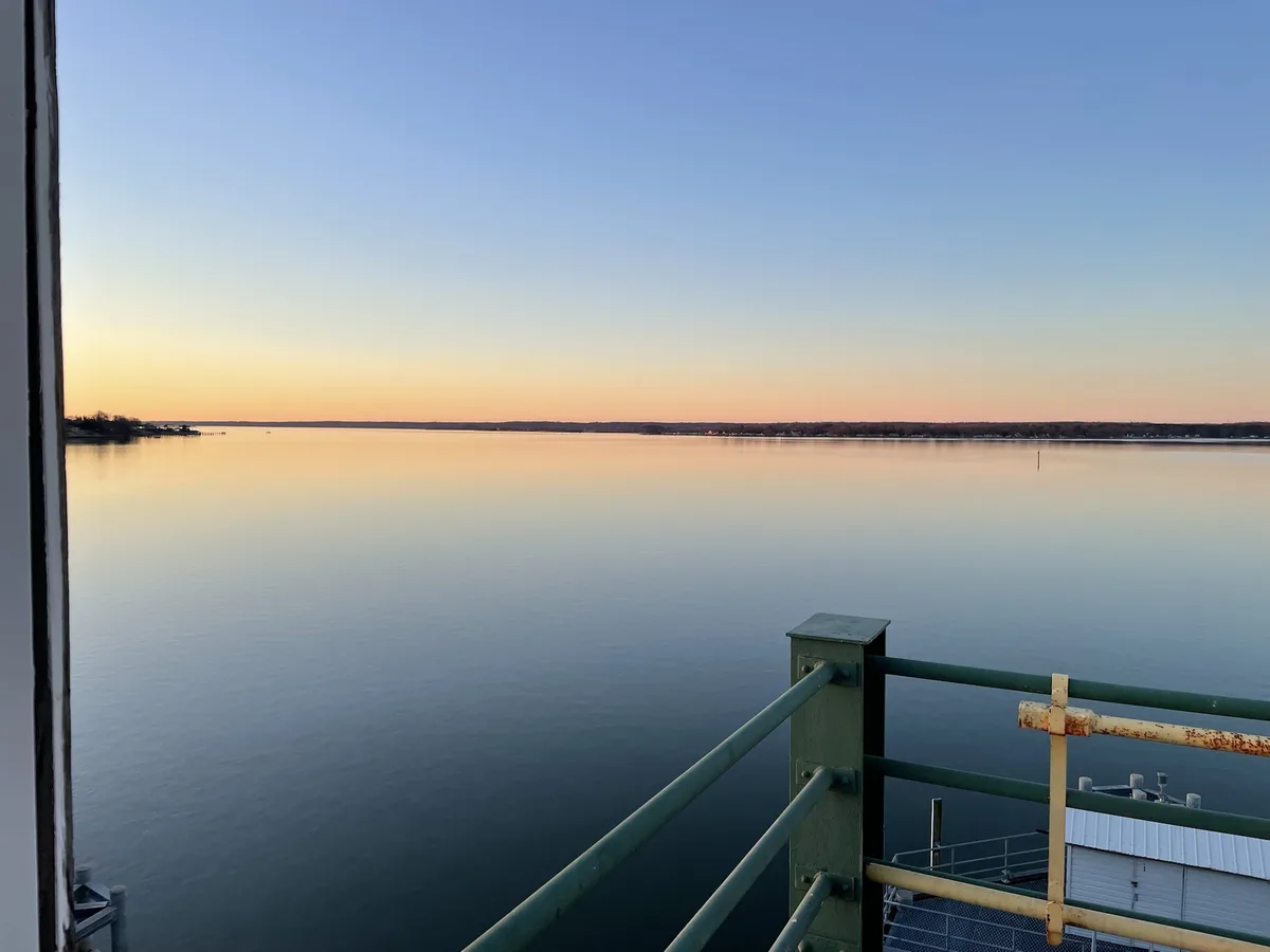 A serene photograph of a calm body of water at sunset, viewed from a railing on a dock or pier. The sky transitions from…