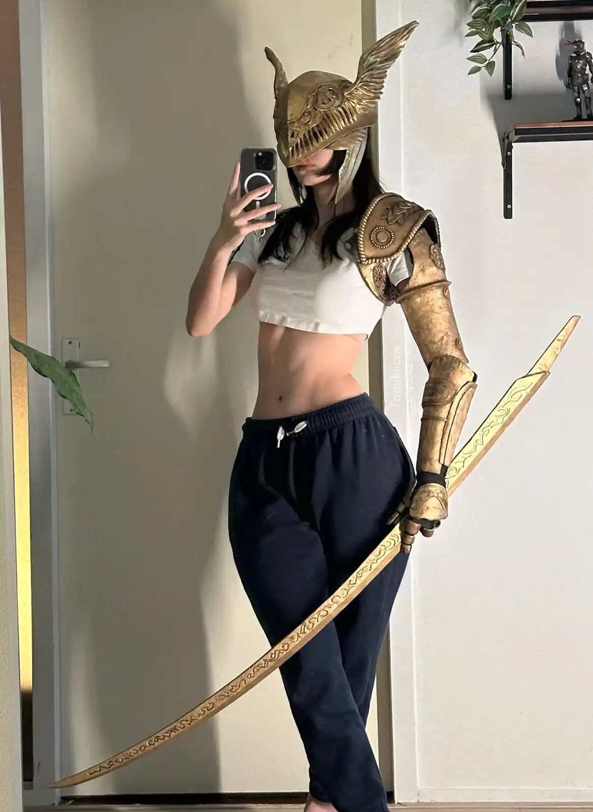 A person in a gold knight helmet and armor takes a mirror selfie while holding a large ornate sword, wearing casual clothes…