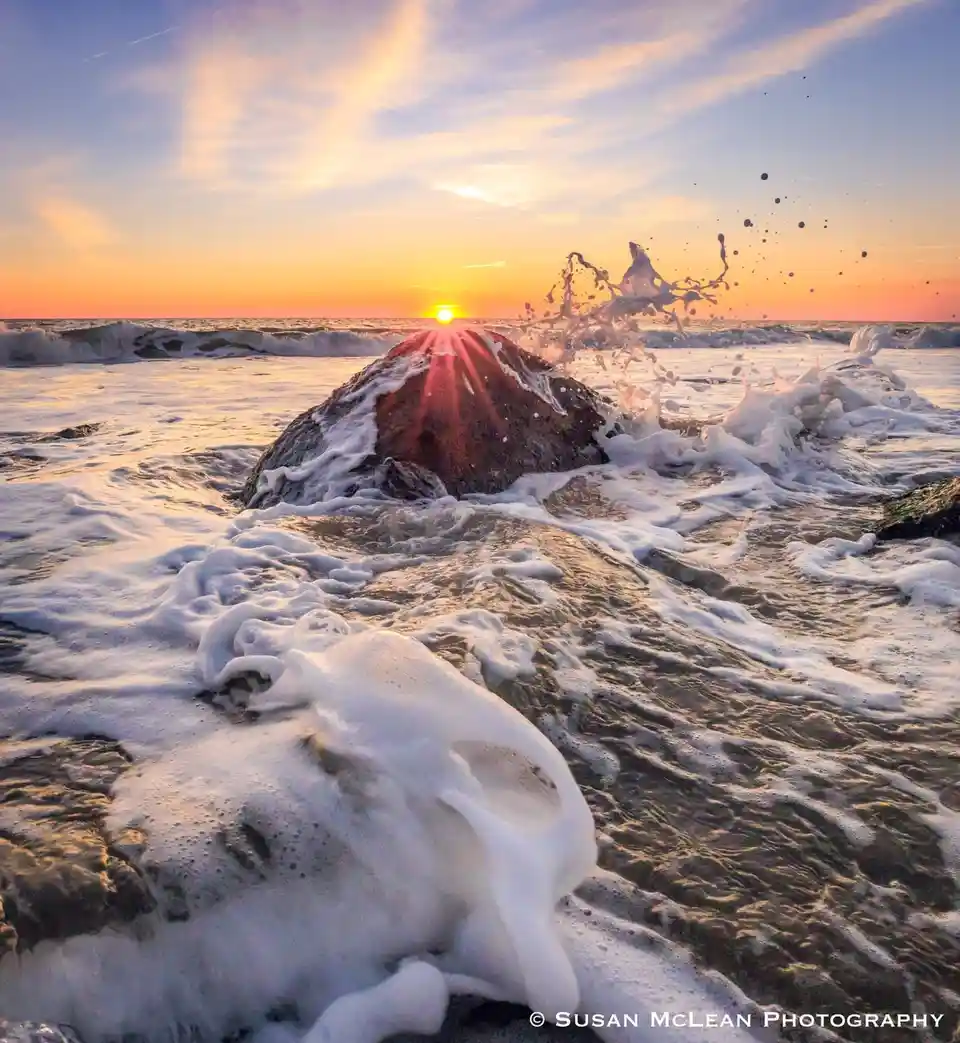 A serene sunset photograph shows waves crashing against rocks, with the sun setting behind a central rock, casting golden…