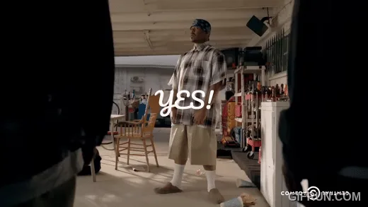 The image shows a man in a plaid shirt and khaki shorts standing in a workshop or garage, looking upward with an excited…