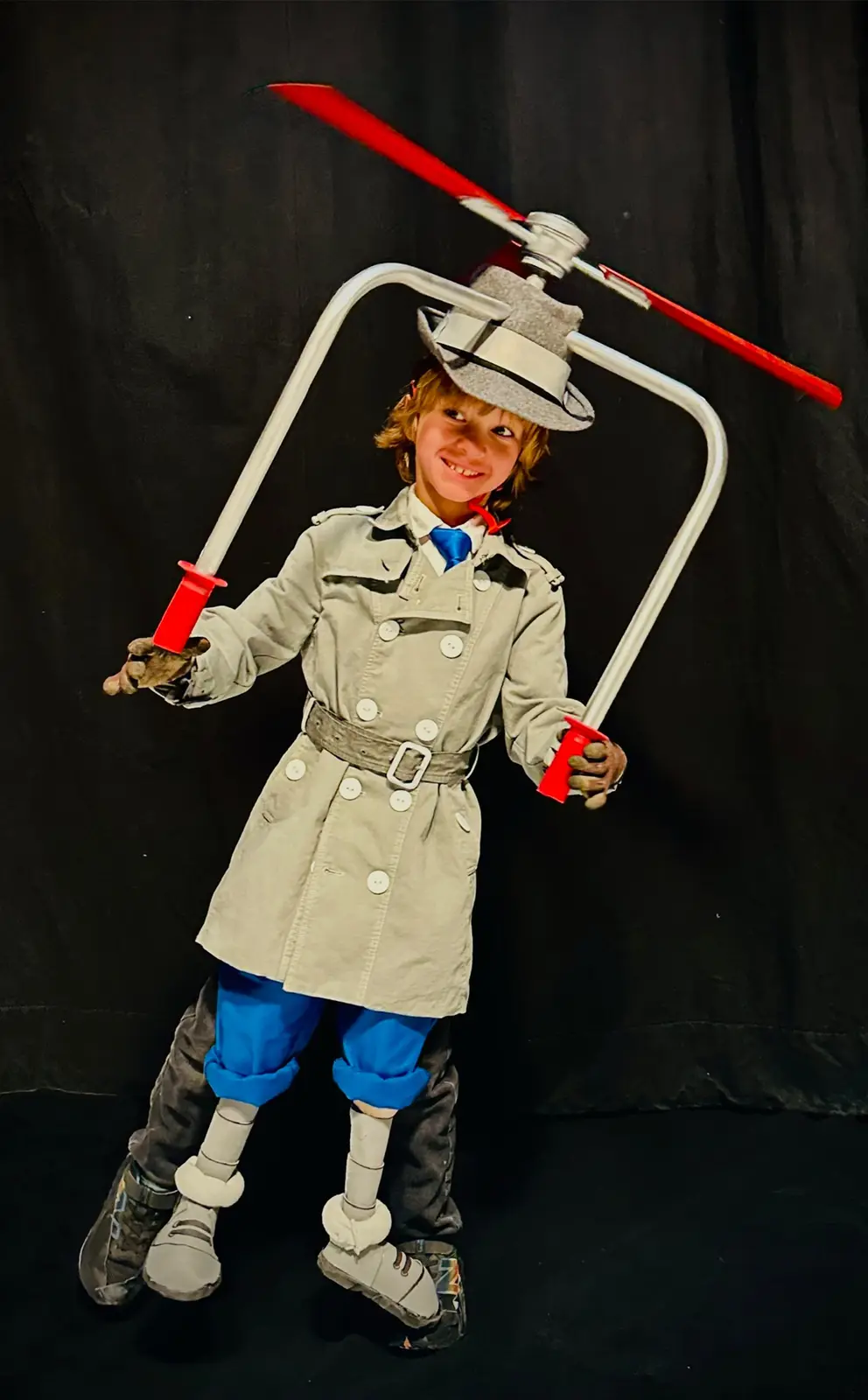 A young child dressed in a beige trench coat, blue pants, and a grey fedora stands against a black backdrop, smiling. A red…