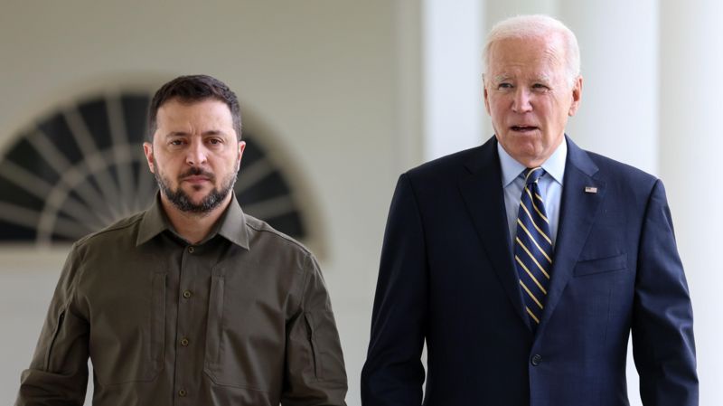 Two men stand side by side in a formal setting; the man on the left wears a brown button-up shirt and has a beard, while the…