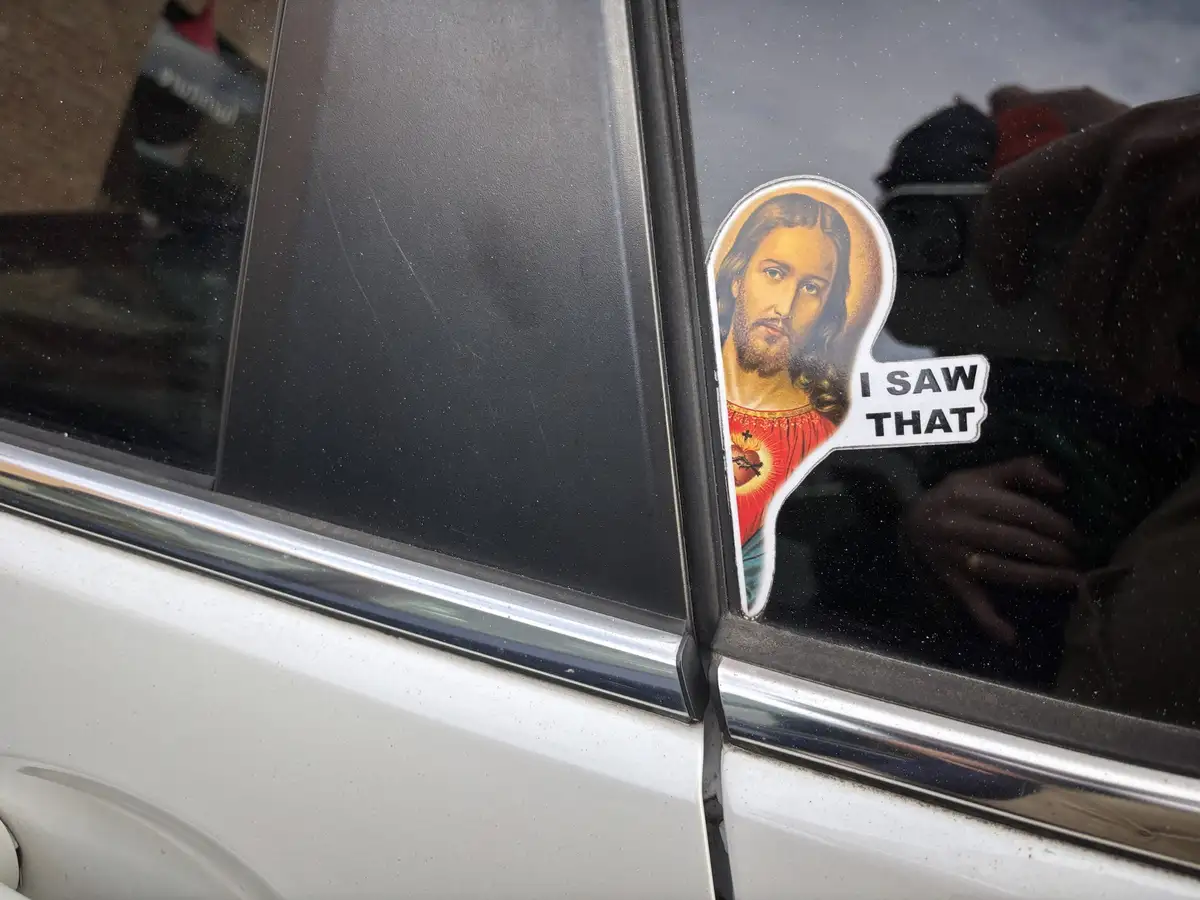 A close-up of a car window shows a religious sticker depicting Jesus Christ with a halo and sacred heart, next to a speech…