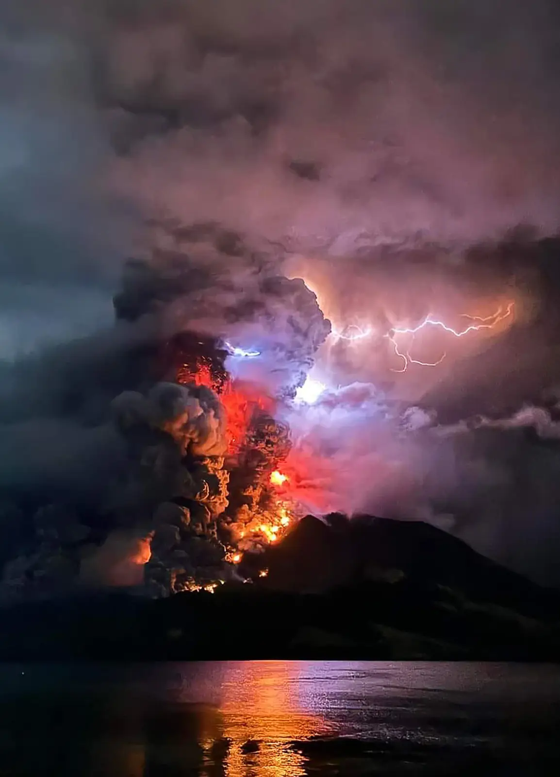 A dramatic volcanic eruption at night, with a massive plume of dark smoke and glowing red lava shooting into the sky…