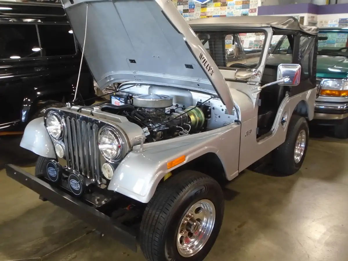 A silver Jeep Wrangler with its hood open, revealing a custom engine, is parked indoors with other vehicles nearby. The word…