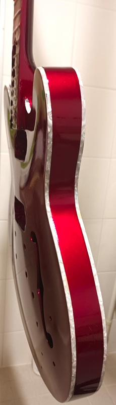 A glossy red electric guitar with a silver binding is shown standing upright against a white tiled wall, its body featuring…