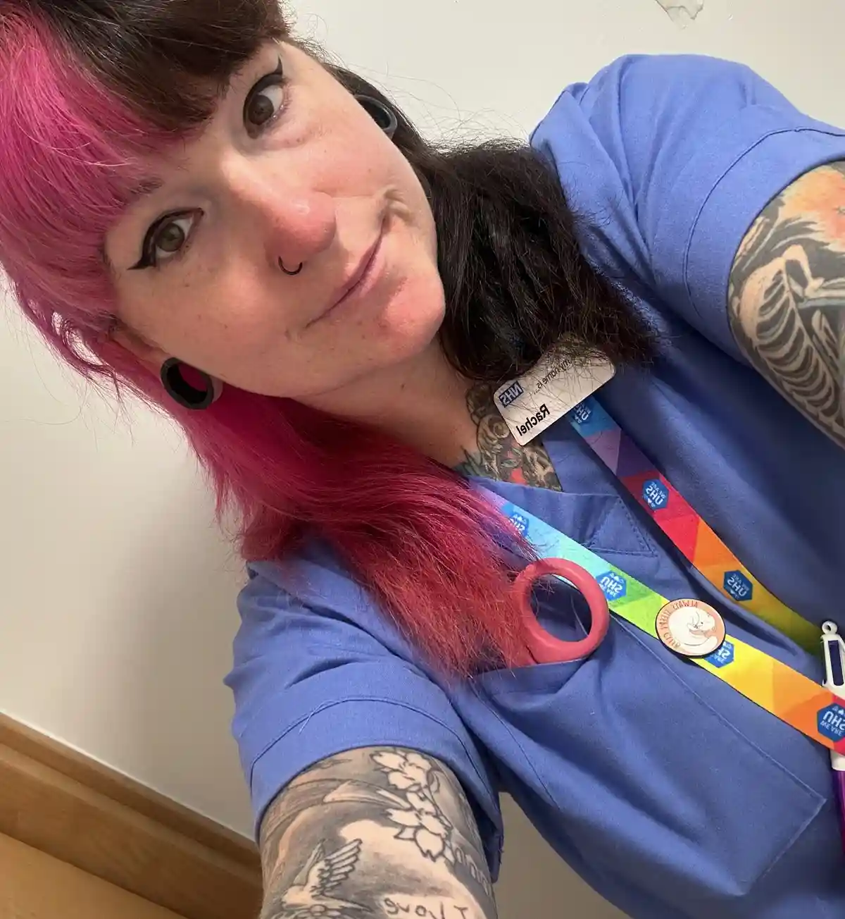 A person with pink and black hair, multiple tattoos on both arms, and a nose ring is wearing blue scrubs and an NHS ID…