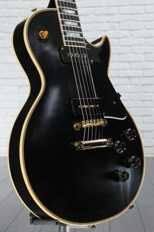 A black electric guitar with gold trim and skull-shaped knobs is shown against a white brick wall. The overlaid text reads…
