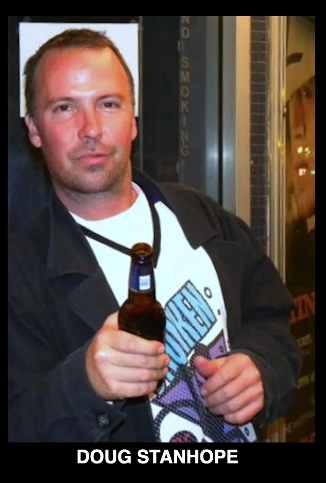 A man with short hair and a slight beard, wearing a black jacket over a white t-shirt with a colorful graphic, holds a brown…